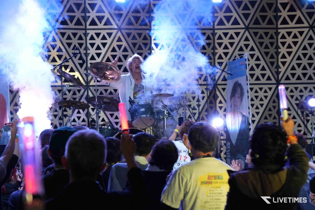 Drummer performing on stage amid blue smoke with a geometric backdrop, audience waving glow sticks in the foreground.