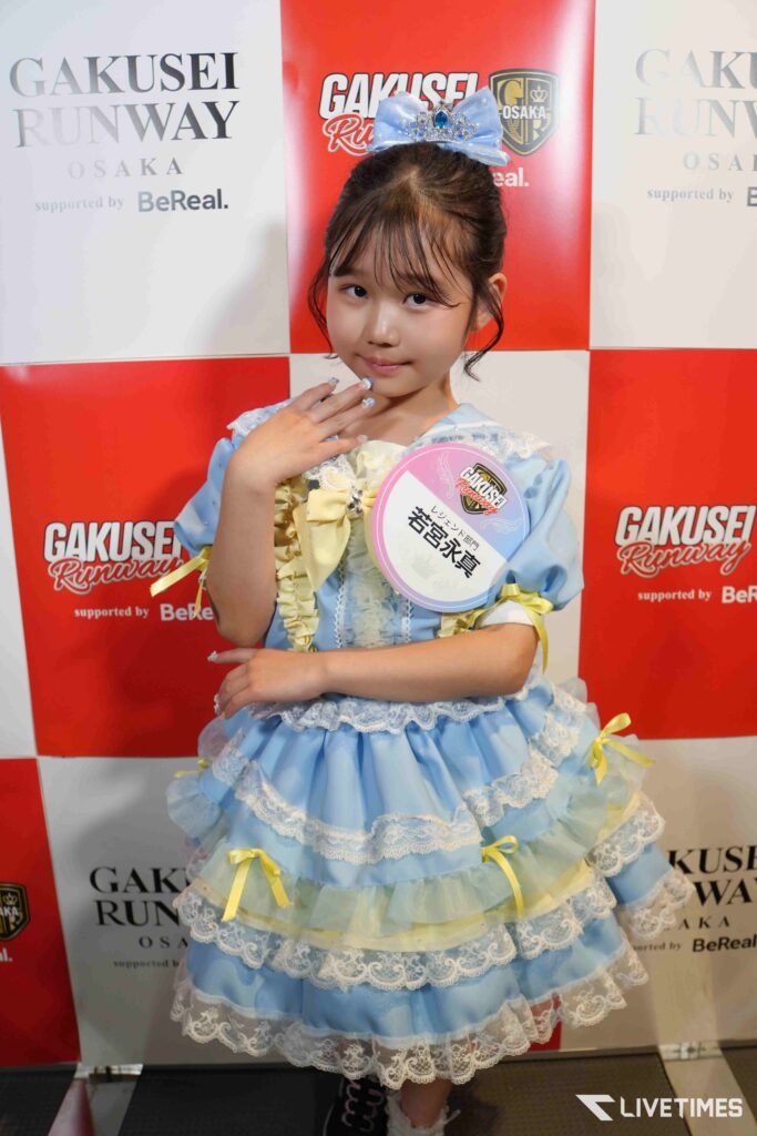 Young girl in a blue frilly dress and tiara posing at a Gakusei Runway backdrop, with a round badge on her chest.