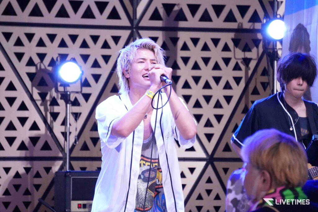 Male singer with bleached hair sings into a microphone on stage, wearing a white shirt, geometric backdrop behind him.