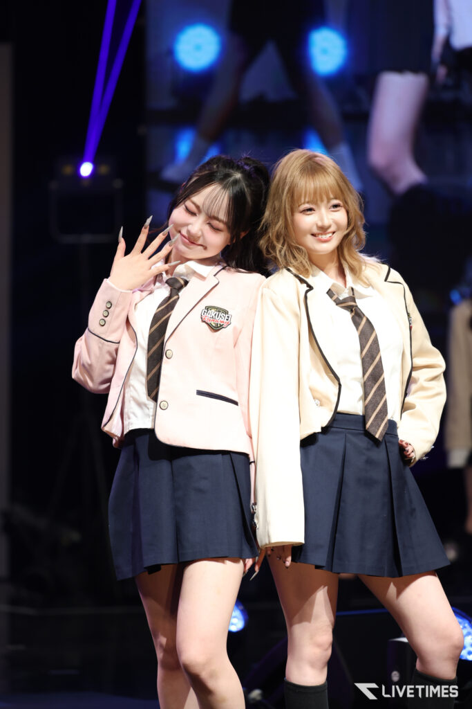 Two young women in matching school uniforms posing on stage, smiling at the camera.