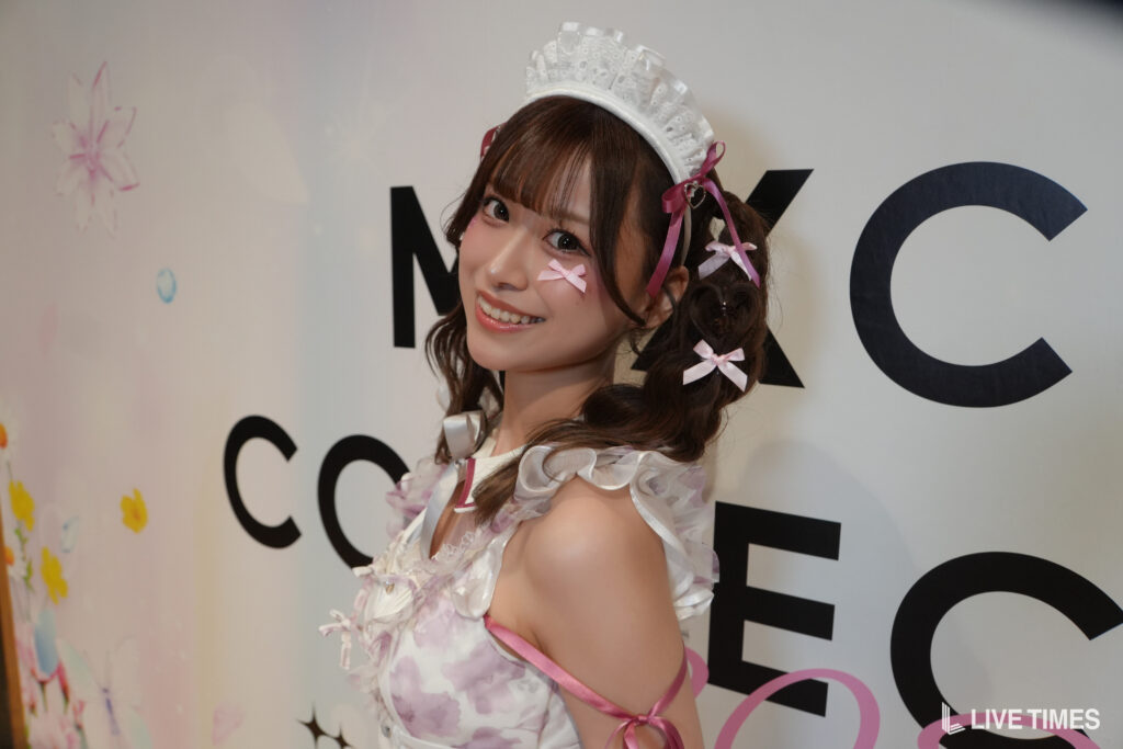 Young woman in a white and pink maid cosplay, bow accents, smiling at the camera.