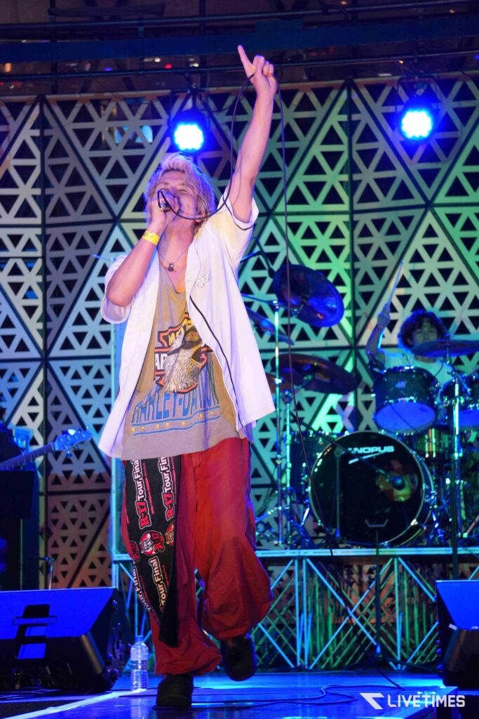 Male singer on stage raising his right arm while singing into a microphone, with a geometric patterned backdrop and drummer in the background