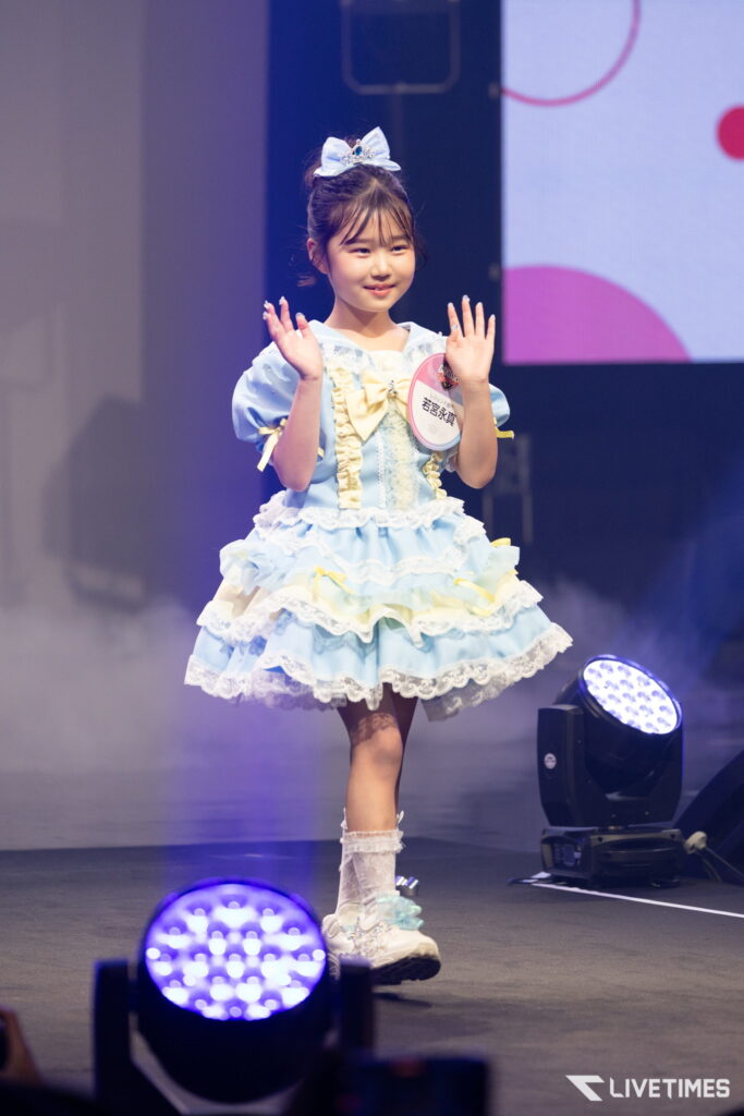 Young girl in a pastel blue frilly dress waves on stage, bow in hair, stage lights in the background.
