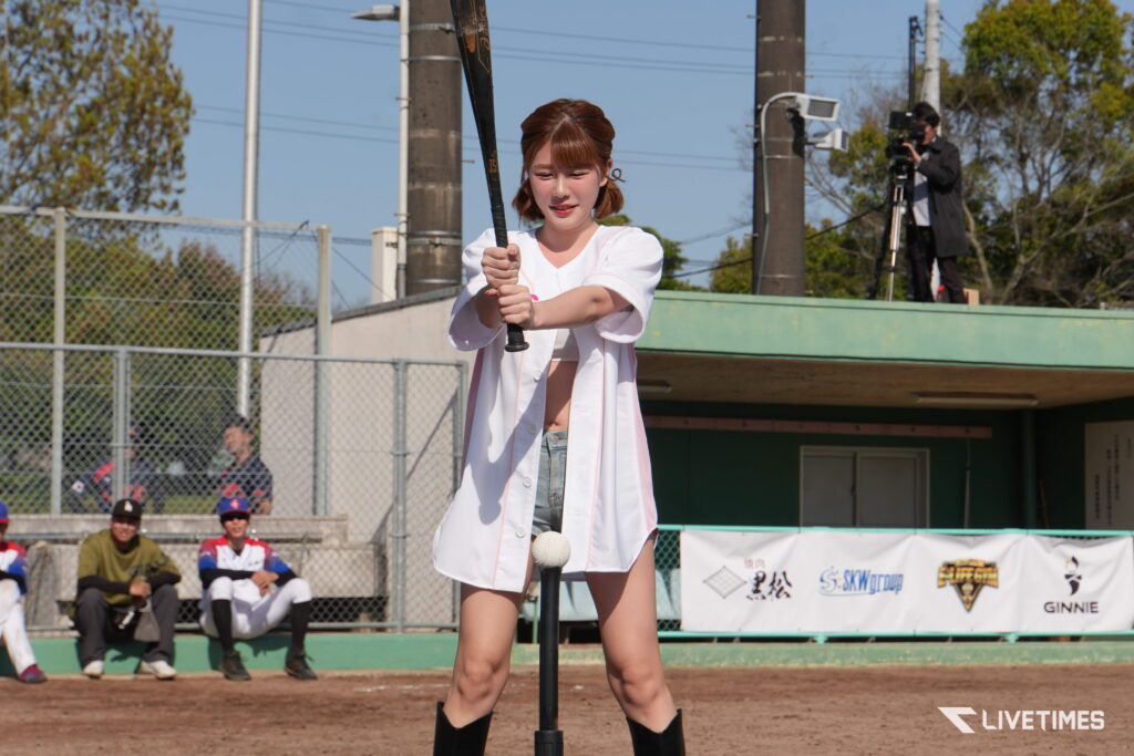 Young woman in a white baseball jersey swings a bat at home plate on a dirt field with a cameraman in the background.