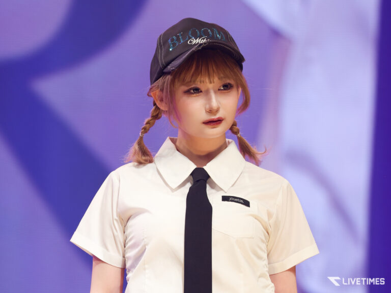 Young woman on stage wearing a white shirt and black tie, with two braided pigtails and a cap reading BLOOM, against a purple backdrop.