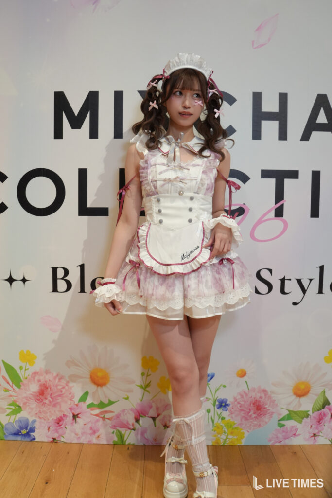 Girl in pastel pink-and-white Lolita-style maid costume with ribbons, posing against a floral backdrop.
