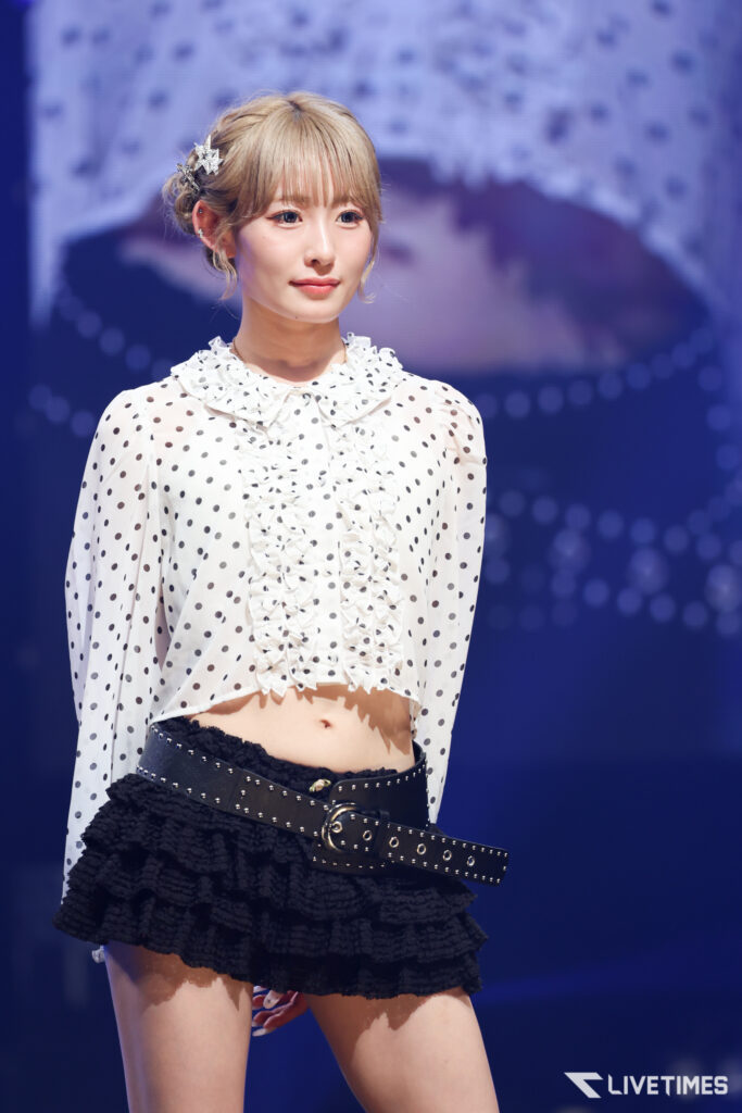 Stage performer wearing a white polka-dot blouse with ruffles, a black studded belt, and a layered black skirt, blue-lit backdrop behind her.