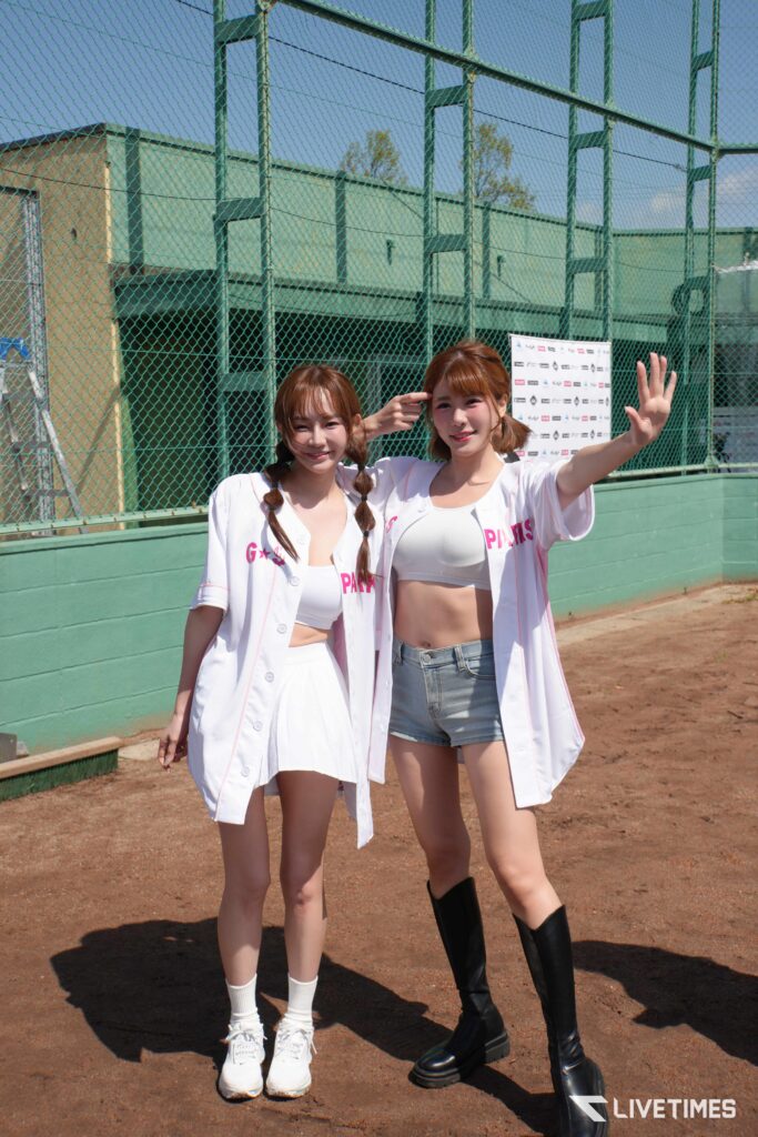 Two young women in white outfits posing for a photo at an outdoor baseball field behind a chain-link fence; one waves to the camera.