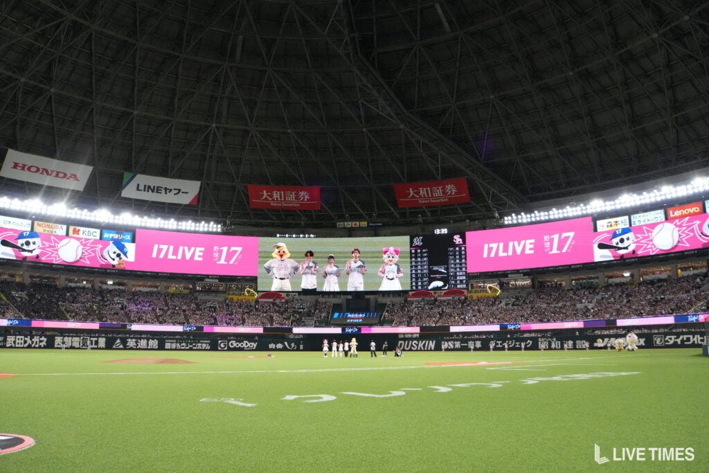 Baseball stadium interior with players on the infield and a large video screen above, surrounded by sponsor banners and bright advertising.