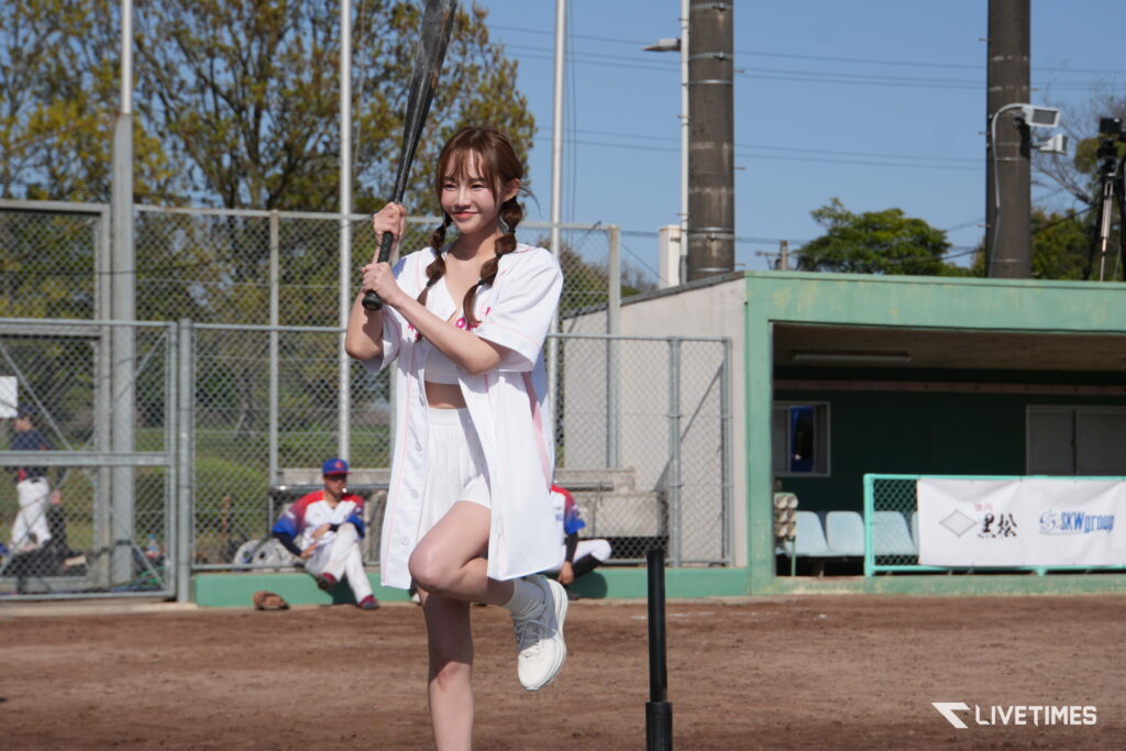 Young woman in a white baseball uniform swinging a bat on a dirt field with a chain-link fence behind her.