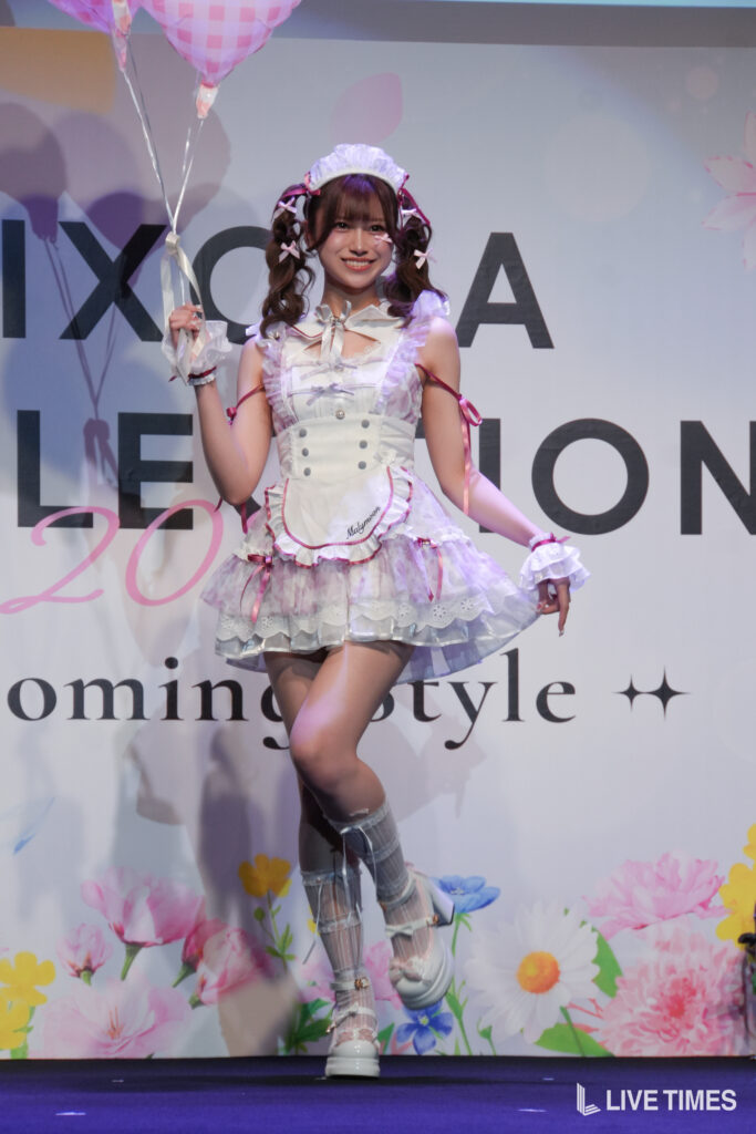 Young woman in a white and pink maid cosplay costume posing on a stage, holding pink balloons with a floral backdrop behind her.