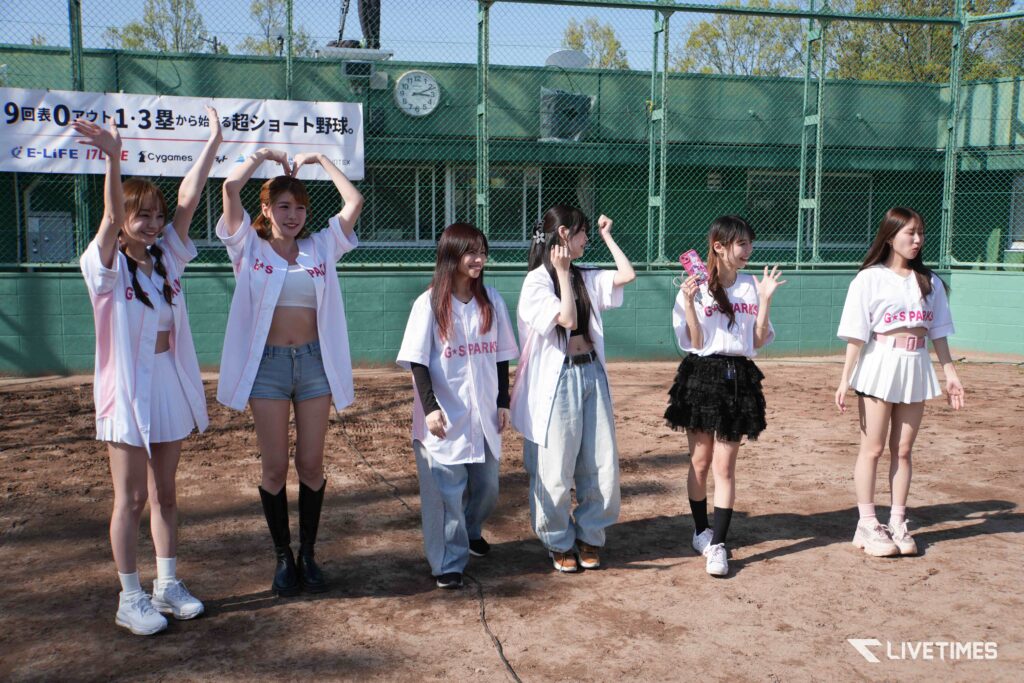 Group of young women in white baseball-style uniforms posing on a dirt field with a chain-link fence and banner behind them.