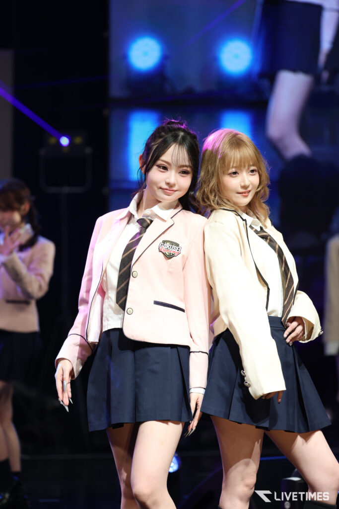 Two performers in pastel blazers and navy skirts pose on a stage with blue spotlights in the background.