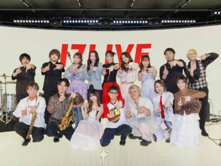 Large group of performers on stage posing for a photo, many pointing or making gestures, with instruments like saxophones visible and a bold red 'LIVE' sign in the background behind them.