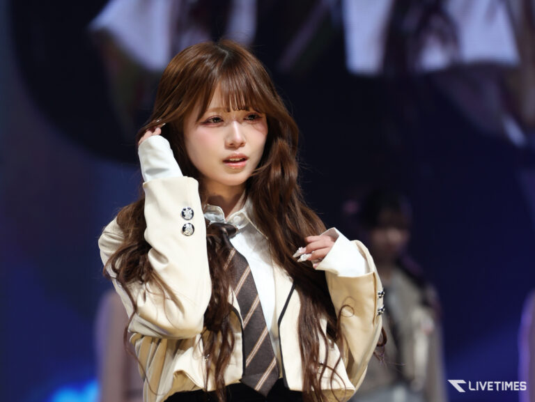 Stage performer adjusting hair, wearing a beige blazer with a brown striped tie, on a dark-lit backdrop.