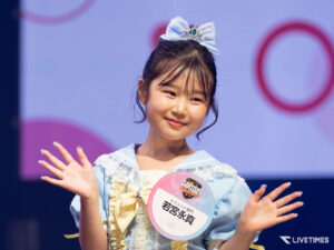 Smiling young girl on stage, wearing a light blue dress, a blue bow tiara, and a round badge on her chest, waving to the audience