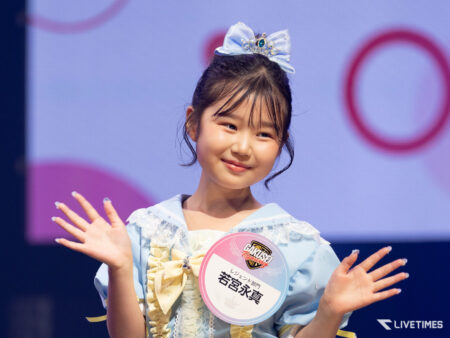 Smiling young girl on stage, wearing a light blue dress, a blue bow tiara, and a round badge on her chest, waving to the audience
