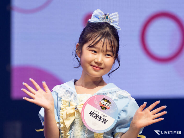 Smiling young girl on stage, wearing a light blue dress, a blue bow tiara, and a round badge on her chest, waving to the audience