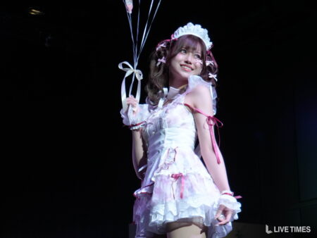 A smiling woman in a white frilly maid outfit on stage, holding white ribbons with blue-tinted background balloons visible to the left.
