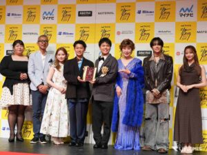 Group of well-dressed people posing on a stage, some holding an award plaque, in front of a yellow‑and‑white branded backdrop.