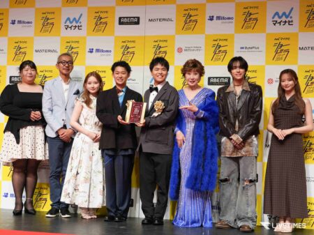 Group of well-dressed people posing on a stage, some holding an award plaque, in front of a yellow‑and‑white branded backdrop.