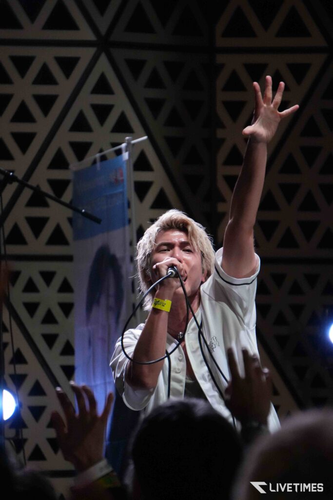 Performer with blond hair singing into a microphone on stage, right arm raised among cheering fans, geometric backdrop behind.
