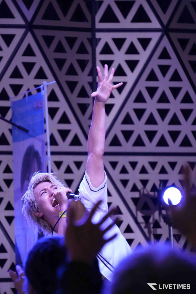 Singer on stage holds a microphone and raises a hand toward the audience under purple lights, with a geometric patterned backdrop behind.