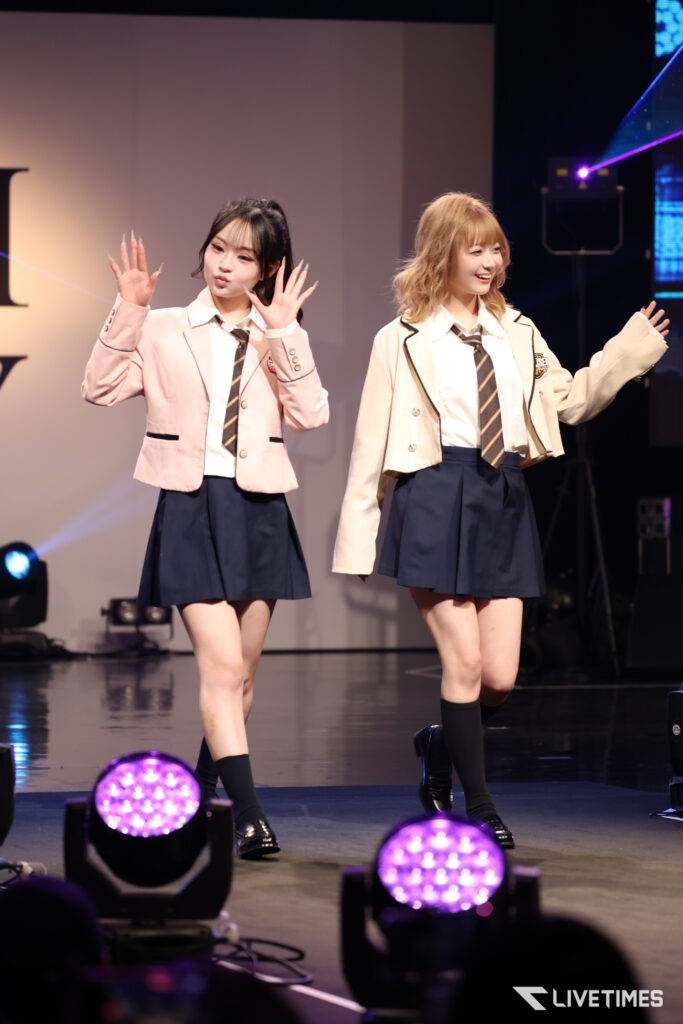 Two young women in school uniform-style outfits perform on stage, waving to the audience.