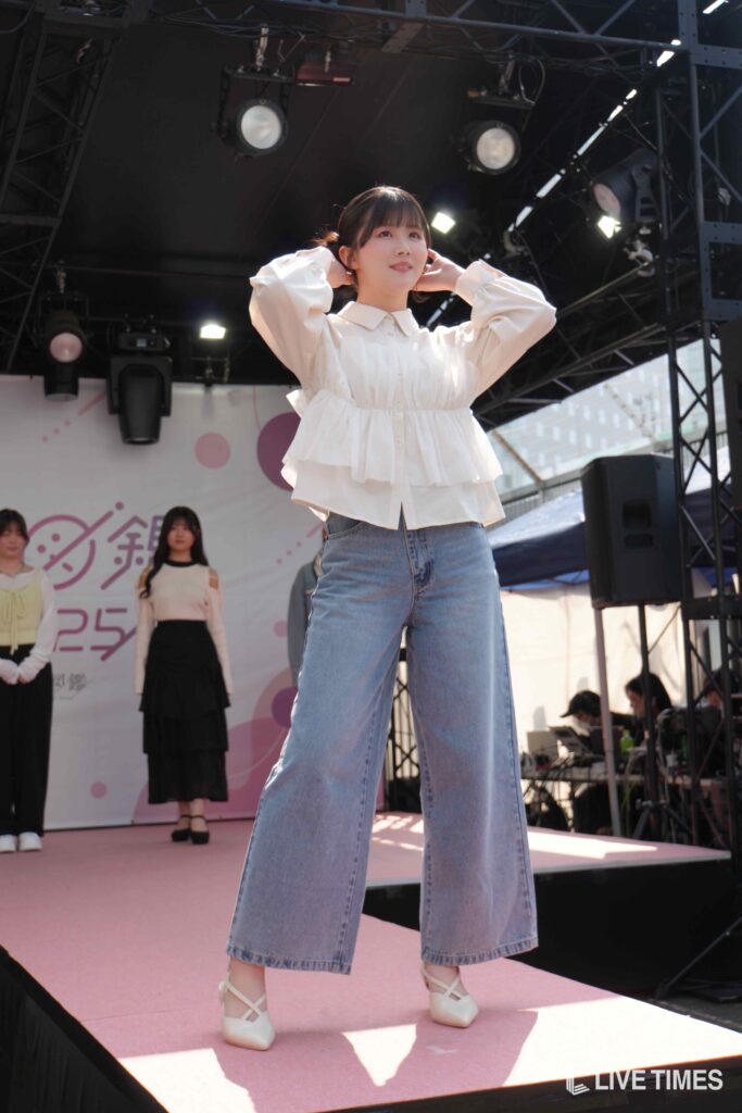 Young woman on a stage posing with hands behind her head, wearing a white blouse and wide-leg jeans, stage lights in the background.
