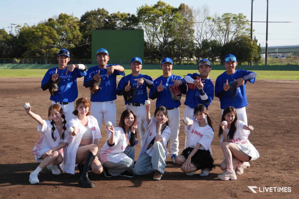 Baseball players in blue uniforms posing with cheerleaders on a dirt field, smiling for a team photo on the diamond.