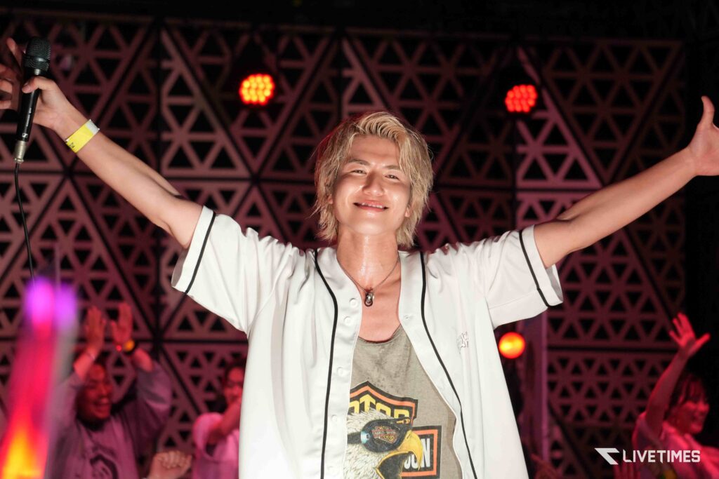 Joyful performer on stage with arms raised, wearing a white shirt, under red stage lights.