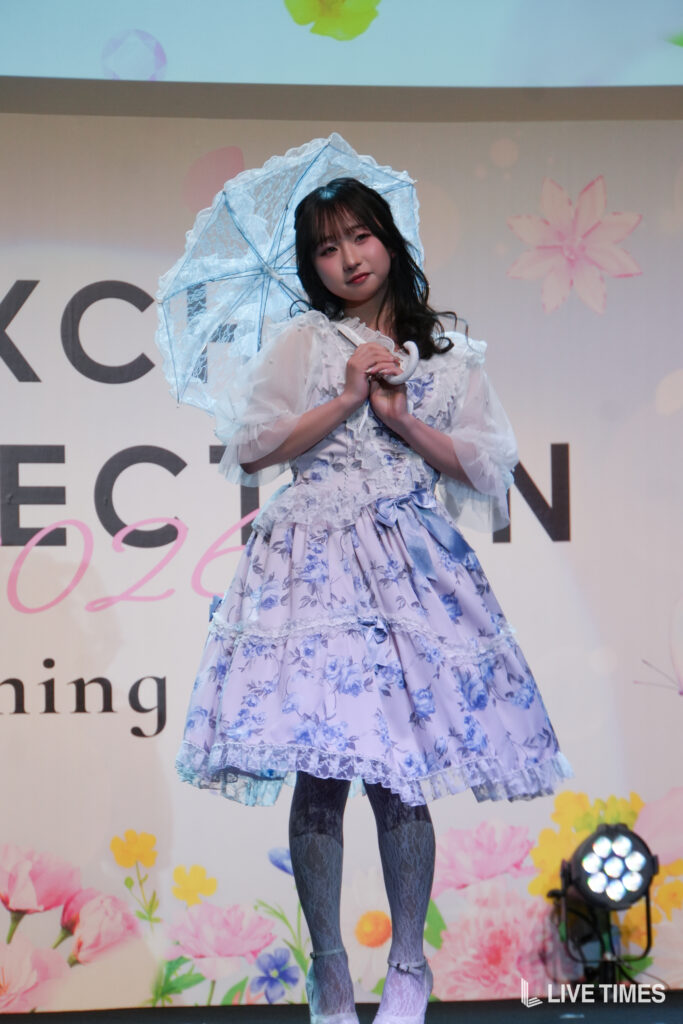 Young woman in a lavender floral Victorian-style dress holding a lace parasol on stage.