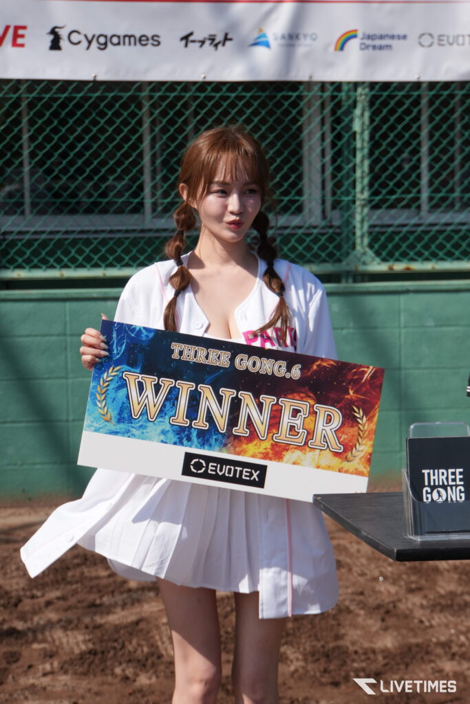 Young woman with braided hair in a white outfit holding a colorful WINNER sign at an outdoor event.