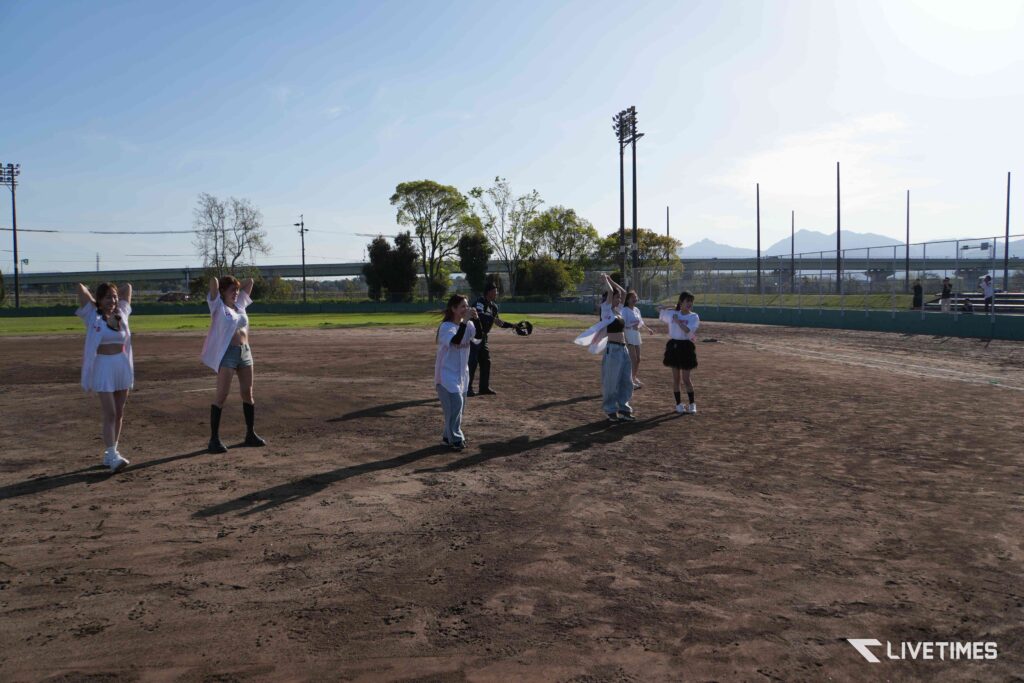 Group of young women in white outfits standing on a dirt baseball field, warming up on a sunny day with a chain-link fence and stands in the background.