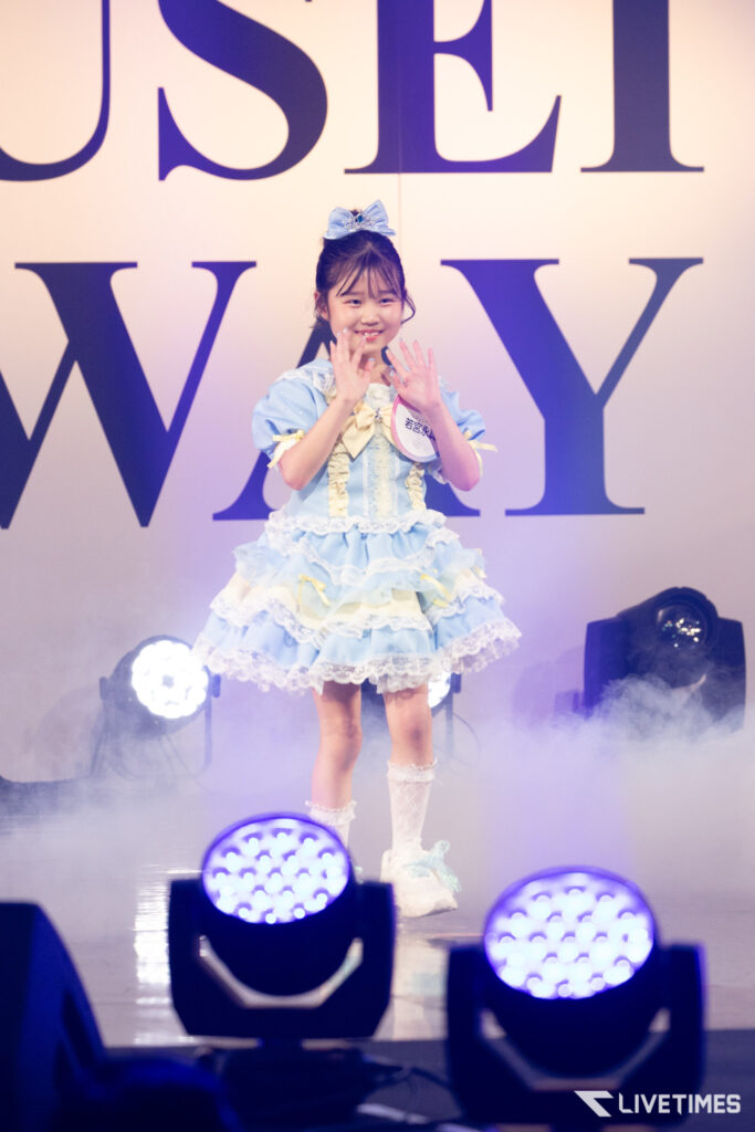 Young girl in a light blue frilly dress waves on stage with fog and large purple letters behind her.