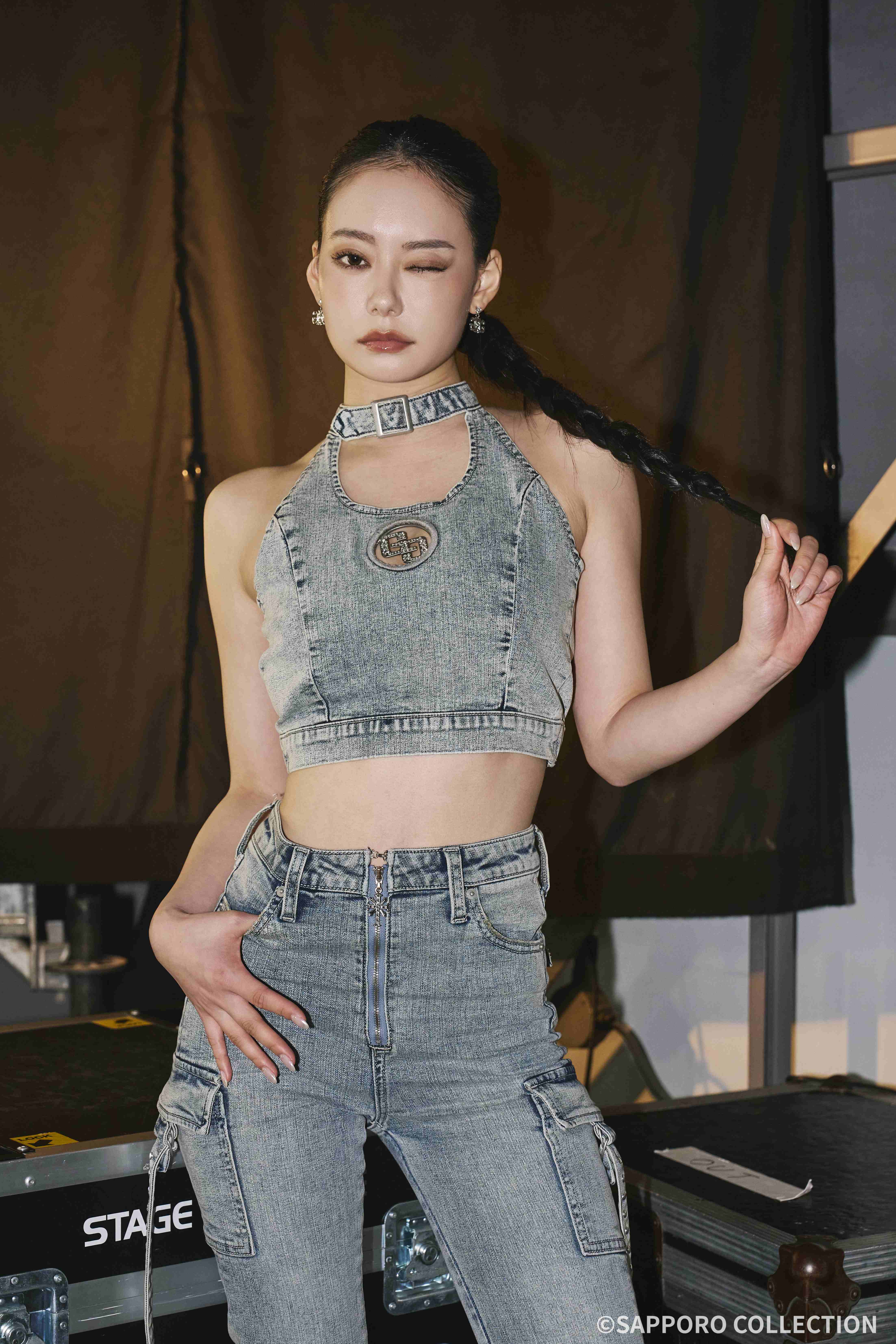 Woman in a denim halter crop top and jeans, winking while holding a braided ponytail beside stage equipment.