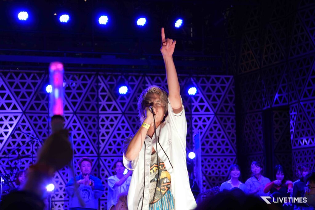 Singer on stage raising one arm with a finger pointing up, performing under blue lights with a cheering crowd in the background.
