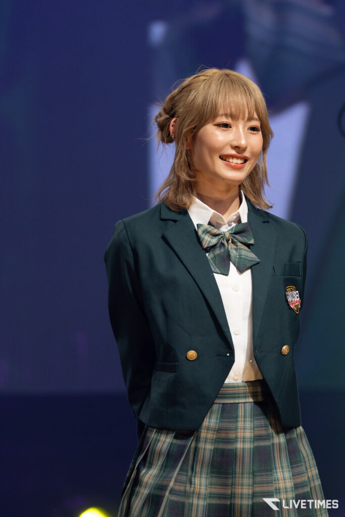 Young woman in a dark green school blazer and plaid skirt, smiling on stage with a bow-tie outfit.