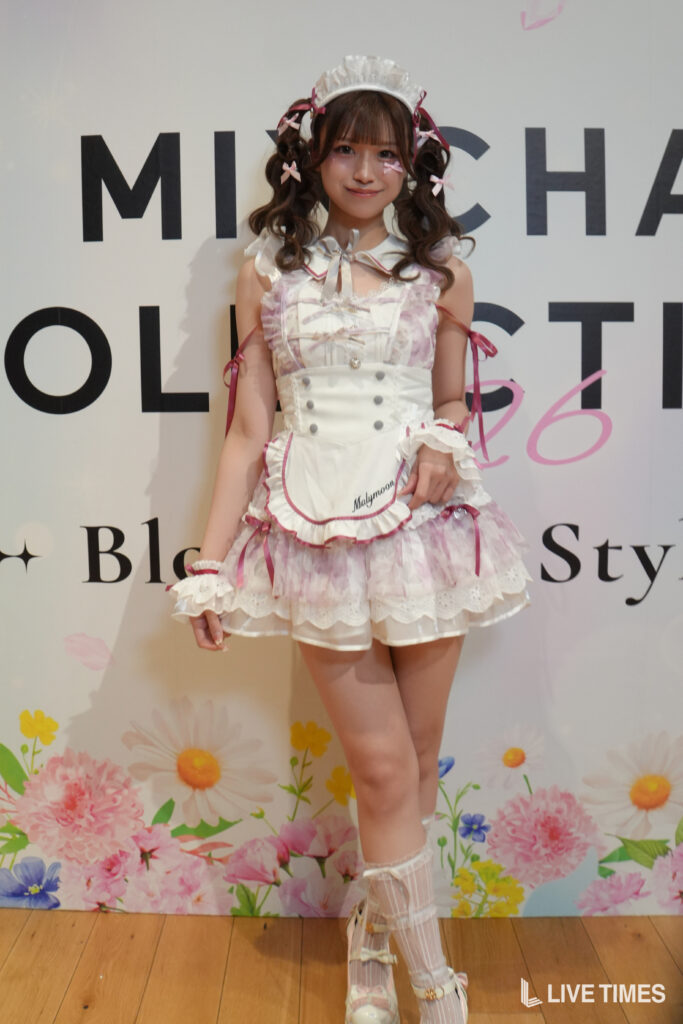 Young woman in a pastel pink and white frilly maid outfit posing on a floral-stage backdrop.