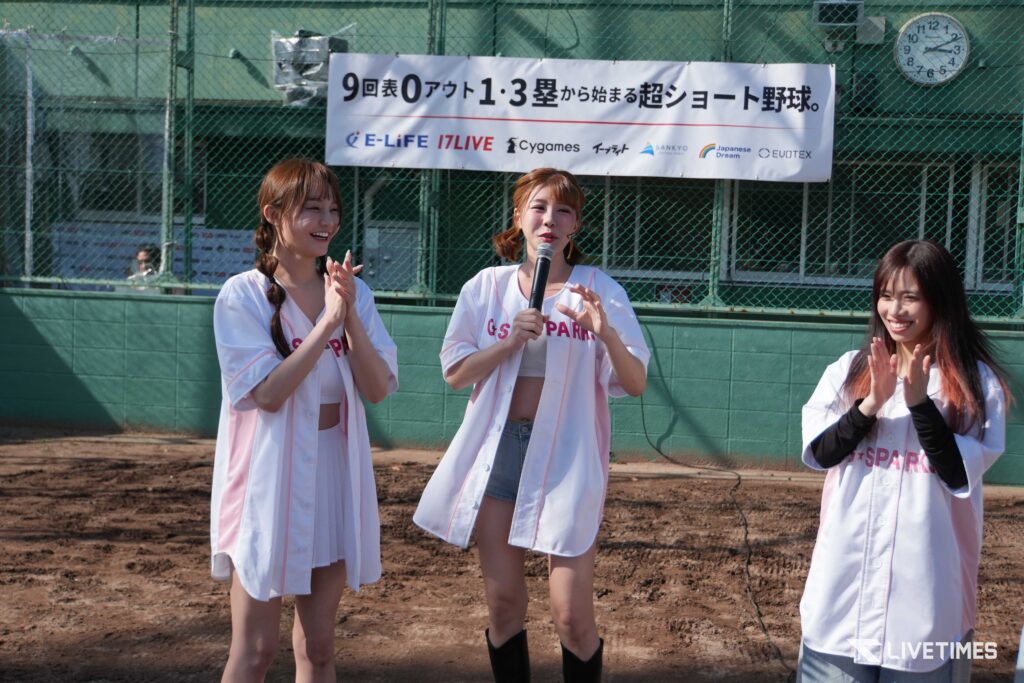 Three young women in white-pink baseball uniforms clap and smile on a dirt field, one holding a microphone with a sponsor banner behind them.