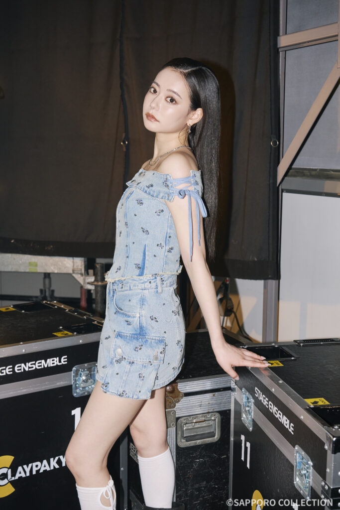 Young woman in a light denim off-shoulder top and skirt, standing backstage beside black gear cases marked Stage Ensemble, looking toward the camera with a calm expression.