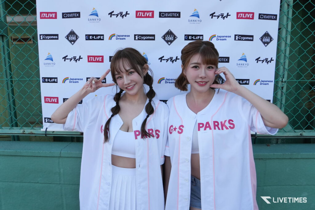 Two young women in white outfits pose for a photo, each making a peace sign in front of a sponsor backdrop with logos like SANKYO, Cygames, and LIVETIME.