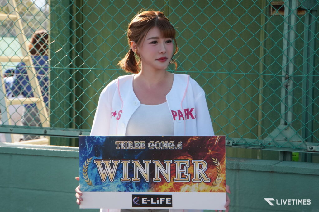 Young woman standing behind a sign that reads 'WINNER' at a game booth, with a green chain-link fence in the background.