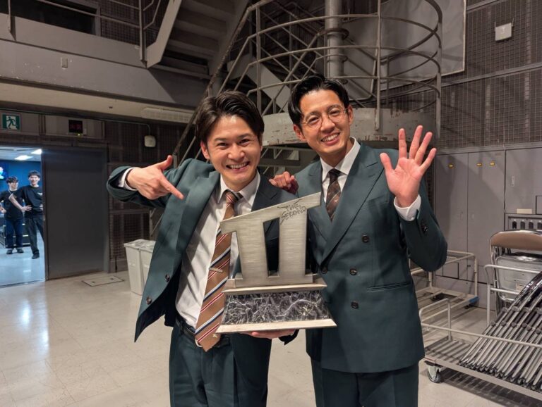 Two men in teal suits smile and pose with a large silver trophy behind them in a backstage area, one pointing at the prize and the other waving.