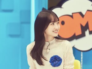 Young woman with long dark hair smiling, wearing a cream knit sweater with a blue pom-pom badge, in a colorful studio set.