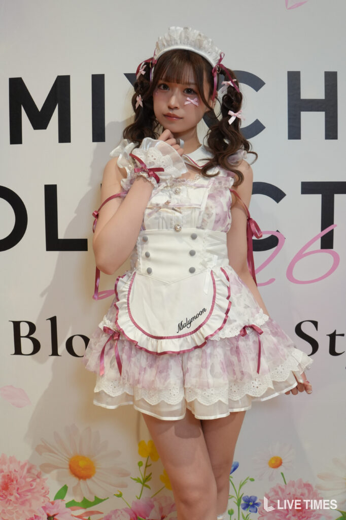 Young girl in a white and pink frilly maid costume, with headband and ribbons, posing with her hand near her chin at an event venue.