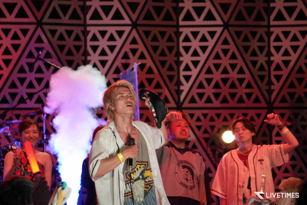 Lead singer with blond hair in a white shirt holds a microphone on stage, surrounded by cheering fans and a pink-blue fog with a geometric triangular backdrop.