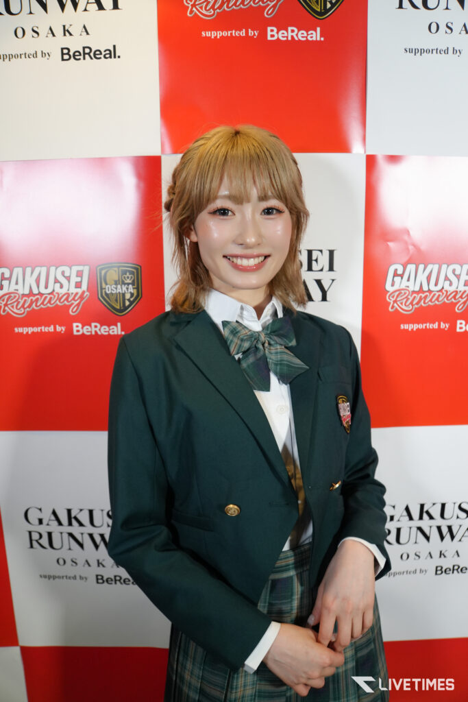 Smiling woman in a dark green blazer and plaid tie posing in front of a red-and-white event backdrop with logos.