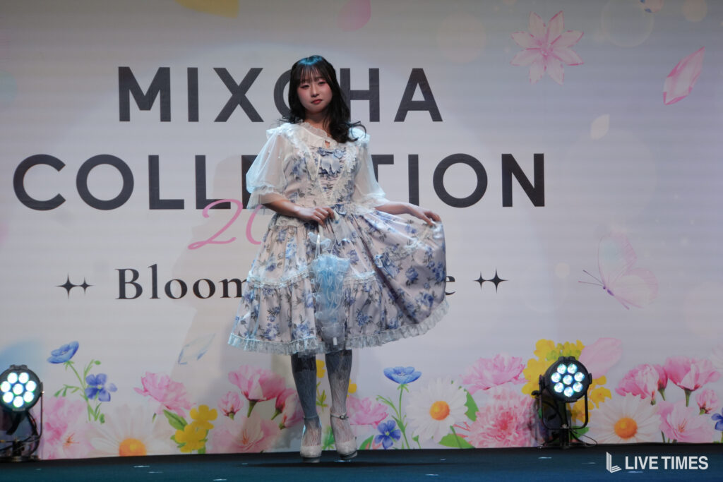Female model in a blue floral, frilly dress posing on a fashion show runway with a MIXCHIA COLLECTION backdrop.