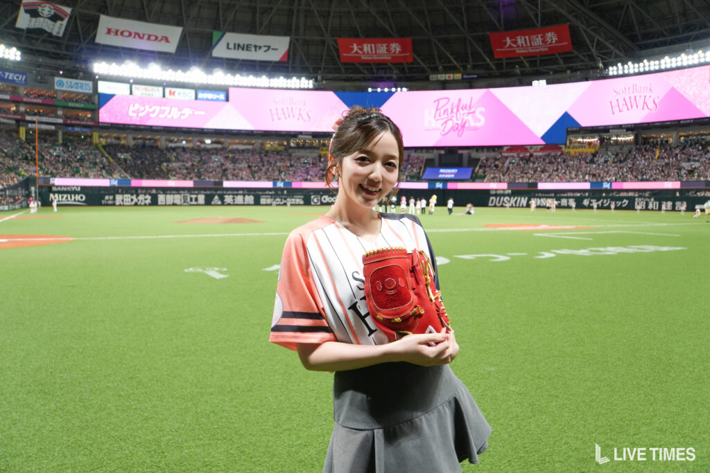 Smiling woman in a baseball jersey holds a red glove on the field, with large pink banners and a crowd behind her.